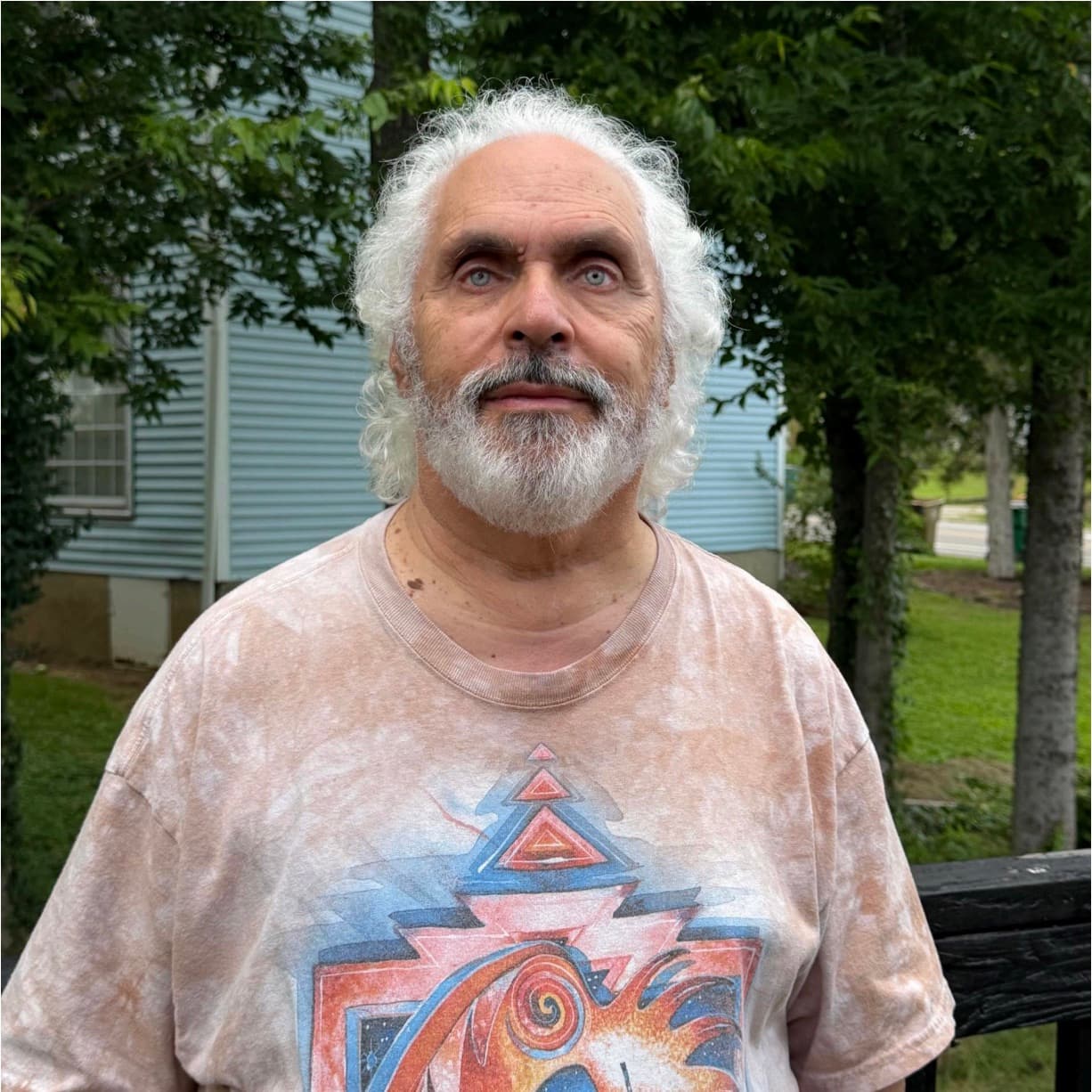 Ross Winetsky standing outdoors in front of trees and a light blue house, wearing a tie-dye graphic t-shirt.