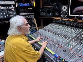 Man with long white hair and beard, wearing a gold shirt, with his hands on a large console with equipment racks and speakers to his side.