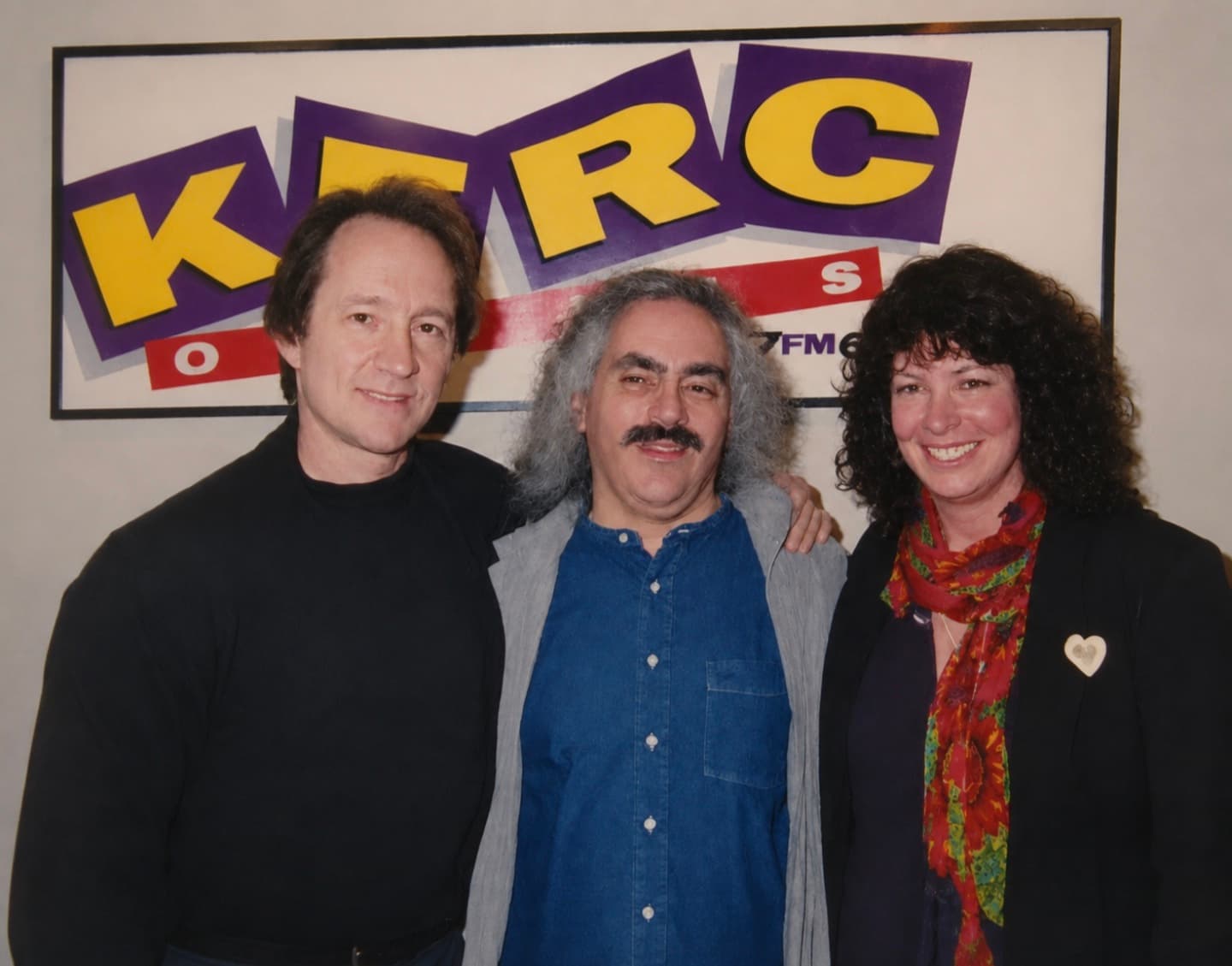 Three people stand arm in arm smiling in front of a large KFRC radio station backdrop. Ross, with long gray curly hair and a mustache, is in the middle wearing a denim shirt and gray vest, flanked by a man in a black turtleneck on the left and a woman with dark curly hair in a black blazer and colorful floral scarf on the right.